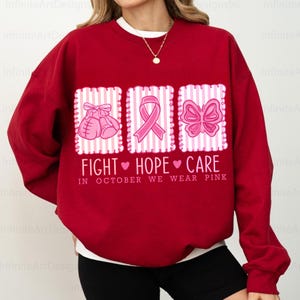 May include: A red sweatshirt with a white crew neck featuring three pink graphic designs. The designs include boxing gloves, a ribbon, and a butterfly, with the words "FIGHT HOPE CARE" and "IN OCTOBER WE WEAR PINK" printed below.