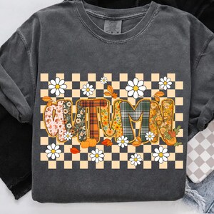 May include: Dark gray sweatshirt with a colorful "AUTUMN" graphic. The letters are filled with floral and plaid patterns, surrounded by daisies, pumpkins, and fall leaves, set against a checkered background.