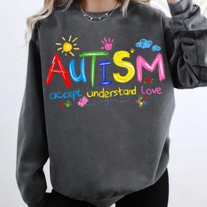 Puede incluir: Sudadera gris con un gr&aacute;fico colorido que dice "AUTISMO" con las palabras "aceptar comprender amar" debajo. El gr&aacute;fico est&aacute; dibujado en un estilo infantil con un sol, nubes y huellas de manos.