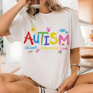 Puede incluir: Camiseta blanca con un dibujo infantil colorido de la palabra "AUTISMO" y las palabras "aceptar comprender amar" debajo.