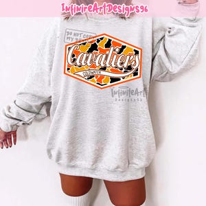 Op de afbeelding: Een lichtgrijs sweatshirt met ronde hals en een centrale zeshoekige afbeelding met een oranje, zwart en wit camouflagepatroon. De tekst "Cavaliers" is in wit cursief over het patroon gedrukt, met "COLDWATER" in kleinere zwarte letters eronder.