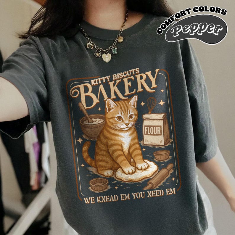 Puede incluir: Camiseta gris oscuro con un gr&aacute;fico que presenta un gato atigrado en un entorno de panader&iacute;a. El dise&ntilde;o incluye las palabras "Kitty Biscuits Bakery" y "We Knead Em You Need Em". La camiseta tambi&eacute;n tiene las palabras "Comfort Colors Pepper" en un c&iacute;rculo.
