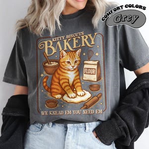 Puede incluir: Camiseta gris oscuro con un gr&aacute;fico de estilo vintage. El gr&aacute;fico presenta un gato atigrado naranja amasando masa, con el texto "Kitty Biscuits Bakery" y "We Knead Em You Need Em". La camiseta tambi&eacute;n tiene las palabras "Comfort Colors" y "Grey".