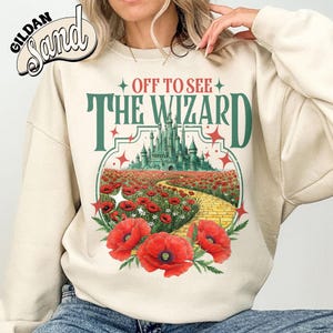 May include: Cream-colored sweatshirt with a graphic featuring the text "OFF TO SEE THE WIZARD" above a scene of a yellow brick road leading to a green castle. Red poppies and stars surround the image.