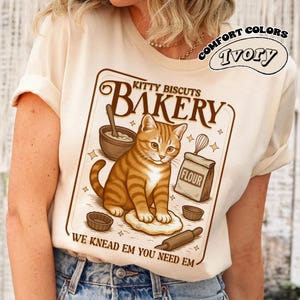 Puede incluir: Camiseta color marfil con una ilustraci&oacute;n de un gato atigrado marr&oacute;n y naranja. El gr&aacute;fico dice "Kitty Biscuits Bakery" con utensilios de panader&iacute;a y el texto "We Knead Em You Need Em". La camiseta tambi&eacute;n tiene las palabras "Comfort Colors" e "Ivory" en un dise&ntilde;o circular.
