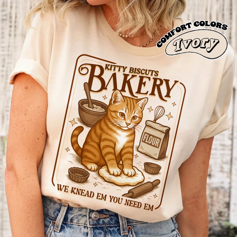 Puede incluir: Camiseta color marfil con una ilustraci&oacute;n de un gato atigrado marr&oacute;n y naranja. El gr&aacute;fico dice "Kitty Biscuits Bakery" con utensilios de panader&iacute;a y el texto "We Knead Em You Need Em". La camiseta tambi&eacute;n tiene las palabras "Comfort Colors" e "Ivory" en un dise&ntilde;o circular.