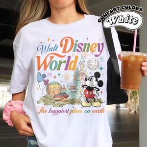 May include: White t-shirt with a colorful Walt Disney World graphic. The design includes Mickey Mouse, a castle, and the phrase "The happiest place on earth." The shirt also has the words "Comfort Colors" and "White" printed on it.