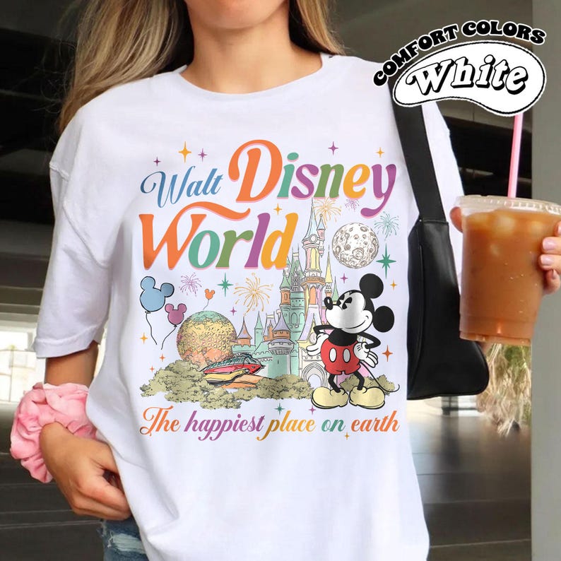 May include: White t-shirt with a colorful Walt Disney World graphic. The design includes Mickey Mouse, a castle, and the phrase "The happiest place on earth." The shirt also has the words "Comfort Colors" and "White" printed on it.