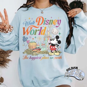 May include: Light blue sweatshirt featuring a colorful Walt Disney World graphic. The design includes Mickey Mouse, a castle, fireworks, and the phrase "The happiest place on earth." The sweatshirt is a Gildan Light Blue.