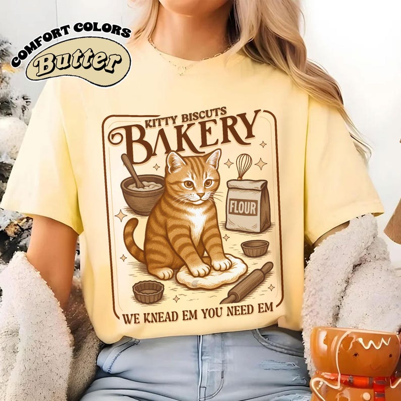 Puede incluir: Camiseta amarilla clara con un dise&ntilde;o de gato atigrado en una panader&iacute;a. El dise&ntilde;o incluye las palabras "KITTY BISCUITS BAKERY" y "WE KNEAD EM YOU NEED EM". La camiseta tambi&eacute;n tiene las palabras "COMFORT COLORS" y "Butter".