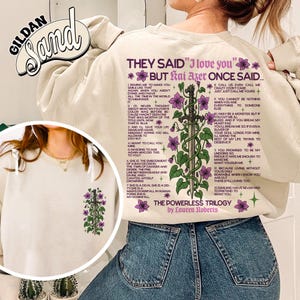 May include: Cream-colored sweatshirt with the text "THEY SAID 'I love you' BUT Kai Azer ONCE SAID..." in purple. The back features a sword design with purple flowers and text. The Gildan Sand brand logo is visible.