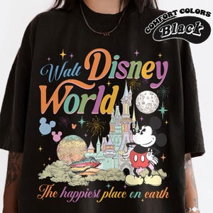 May include: Black t-shirt featuring a colorful graphic of Walt Disney World. The design includes the park's logo, Mickey Mouse, a castle, and the phrase "The happiest place on earth." The shirt is labeled "Comfort Colors Black."