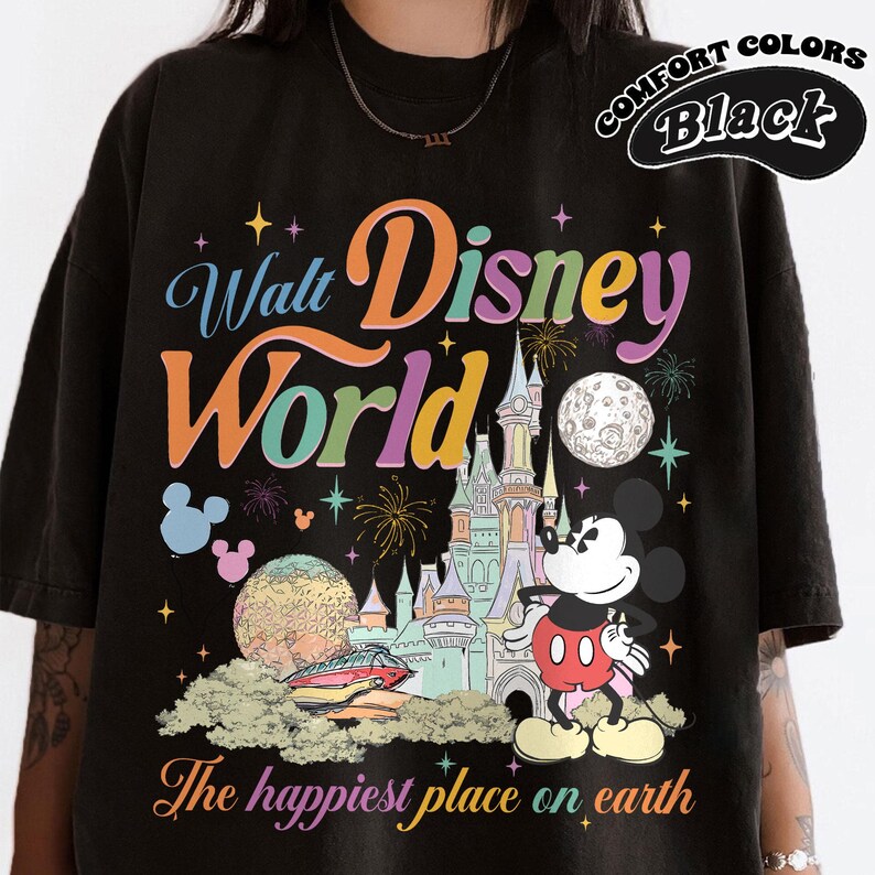 May include: Black t-shirt featuring a colorful graphic of Walt Disney World. The design includes the park's logo, Mickey Mouse, a castle, and the phrase "The happiest place on earth." The shirt is labeled "Comfort Colors Black."