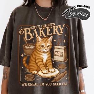 Puede incluir: Camiseta gris oscuro con un gr&aacute;fico de estilo vintage. El gr&aacute;fico presenta un gato atigrado naranja, las palabras "KITTY BISCUITS BAKERY" e ilustraciones relacionadas con la panader&iacute;a. Tambi&eacute;n se incluye el texto "WE KNEAD EM YOU NEED EM".