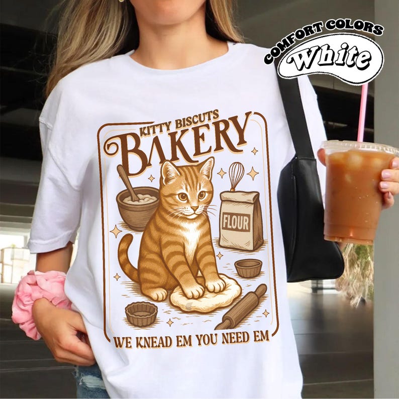 Puede incluir: Camiseta blanca con un gr&aacute;fico marr&oacute;n de un gato en una panader&iacute;a. El dise&ntilde;o incluye el texto "Kitty Biscuits Bakery" y "We Knead Em You Need Em". La camiseta tambi&eacute;n tiene impreso "Comfort Colors".