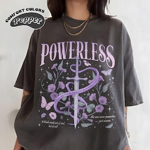 May include: A dark gray t-shirt with a purple and white graphic design featuring a sword, flowers, and the text "POWERLESS". The text below the graphic reads "FOR EVERY GIRL WHO HAS EVER FELT POWERLESS" and "She was never powerless - just unseen".
