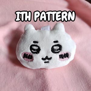 Könnte beinhalten: Ein weißes, plüschiges, katzenförmiges Objekt mit schwarzen Augen, einem Schnurrbart und rosa Wangen. Der Text "ITH PATTERN" wird über der Katze angezeigt. Das Objekt befindet sich auf einem rosa Hintergrund.