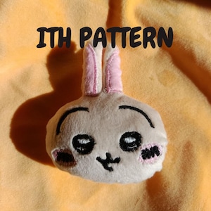 Può includere: Un peluche a forma di coniglietto con corpo grigio chiaro, interno delle orecchie rosa e dettagli del viso ricamati in nero. Le guance hanno accenti rosa. Il testo "ITH PATTERN" è sopra il coniglietto.