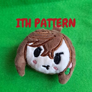 May include: A plush doll in the shape of a cartoon character's head, featuring brown hair, black eyes, and a white face. The doll has a small pink flower and green leaves on the side. The text "ITH PATTERN" is written in red above the doll.