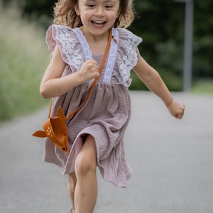 Puede incluir: Una niña pequeña con un vestido morado claro con ribetes de encaje blanco y una corona de flores corre hacia la cámara. Lleva un bolso de cuero marrón con forma de ciervo.