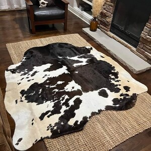 May include: A black and white cowhide rug with a natural, tan-coloured background. The rug is laid out on a woven, natural fibre rug, which is on a hardwood floor.