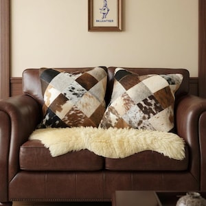 May include: A brown leather sofa with two patchwork cowhide cushions in shades of brown, black, and white. A cream-colored sheepskin rug is draped over the sofa. A framed picture hangs on the wall above the sofa. The picture has the text "BK.LEATHER".