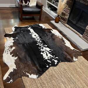 May include: A black and white cowhide rug with brown patches, laid out on a woven rug in a living room setting. The rug is in front of a fireplace with a brown leather armchair and a throw pillow.