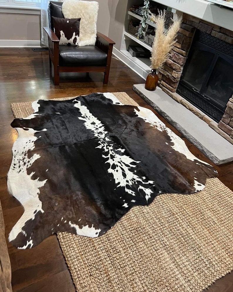 May include: A black and white cowhide rug with brown patches, laid out on a woven rug in a living room setting. The rug is in front of a fireplace with a brown leather armchair and a throw pillow.