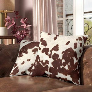 May include: Two square throw pillows featuring a brown and white cowhide print pattern. The pillows are displayed on a brown leather sofa, showcasing a distinct spotted hide texture with irregular dark brown patches against a white background.