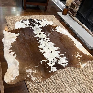 May include: A brown and white cowhide rug with a natural, rustic look. The rug is laid on a woven jute rug, creating a layered look.