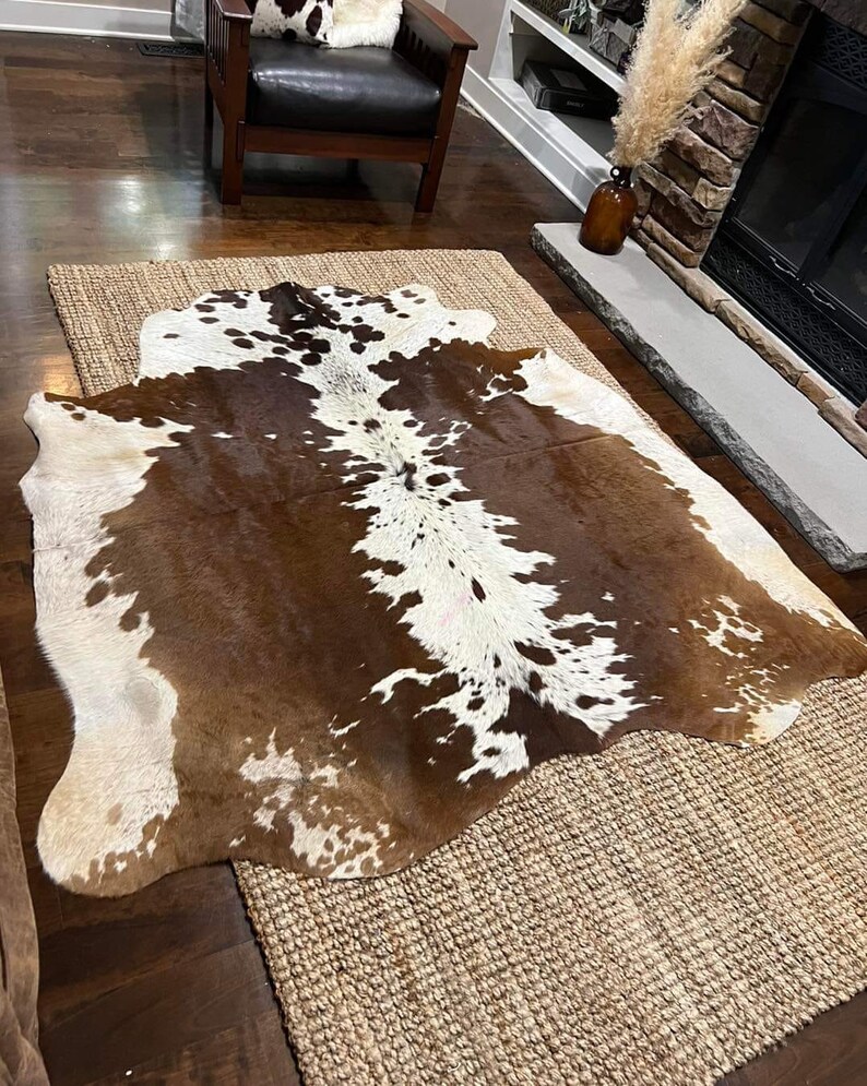 May include: A brown and white cowhide rug with a natural, rustic look. The rug is laid on a woven jute rug, creating a layered look.