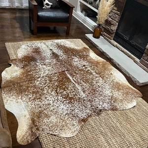 May include: A brown and white cowhide rug with a natural, irregular shape. The rug is laid on top of a woven jute rug.