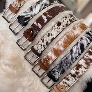 May include: A collection of eight belts featuring various animal print patterns on hair-on hide leather. Each belt includes a silver-toned metal buckle and a leather keeper loop. The patterns range from spotted brown and white to striped black and white.
