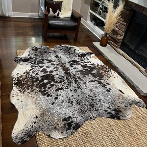 May include: A black and white cowhide rug with a brown and white pattern. The rug is laid out on a woven rug with a light brown colour.