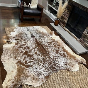 May include: A brown and white cowhide rug with a natural, spotted pattern. The rug is laid out on a woven rug in a living room setting.