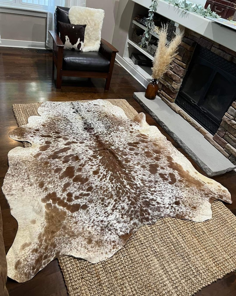 May include: A brown and white cowhide rug with a natural, spotted pattern. The rug is laid out on a woven rug in a living room setting.