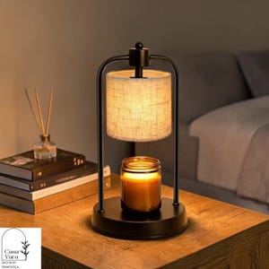 Op de afbeelding: Een zwarte metalen kaarsenwarmer met een cilindrische lampenkap en een kaars in een pot. De warmer staat op een houten oppervlak, met boeken en een diffuser op de achtergrond. De kaars is aangestoken en geeft een warme gloed af.