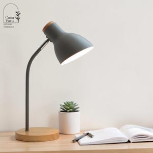 May include: A grey desk lamp with a wooden base and a wooden accent on the lamp head. The lamp is on a light-coloured wooden surface. A small succulent in a white pot, an open notebook, and a pen are also on the surface.