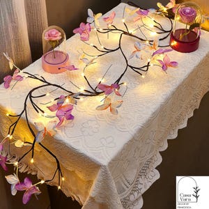 May include: A decorative table setting featuring a butterfly light garland and two glass domes with preserved roses. The garland has brown branches, pink and purple butterflies, and warm white lights. The table is covered with a white lace tablecloth.