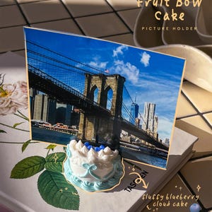 Könnte beinhalten: Ein Bildhalter mit einem Foto der Brooklyn Bridge und einem kleinen Kuchen. Der Kuchen ist weiß mit blauem Zuckerguss und Blaubeeren. Der Text auf dem Bildhalter lautet "Cream Fruit Bow Cake PICTURE HOLDER".
