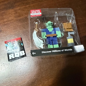 May include: A Roblox action figure set in a clear plastic package. The figure is a green and blue block character with accessories. A small card with the letters "ROB" is also included. The package has text that says "Discover Millions of Worlds."