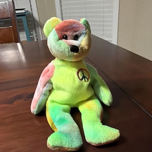 Könnte beinhalten: Ein mehrfarbiger Plüschbär mit einem Friedenssymbol auf der Brust. Der Bär hat eine Mischung aus Regenbogenfarben, darunter Grün, Gelb, Rosa und Rot. Er hat eine schwarze Nase und einen freundlichen Ausdruck, was ihn zu einem fröhlichen und lebendigen Spielzeug macht.