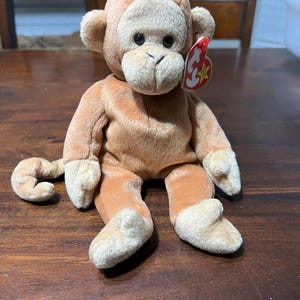 May include: A plush monkey toy with a light brown body and a tan face, ears, hands, and feet. The monkey has black eyes and a red tag attached to its ear. The toy is sitting on a wooden surface.
