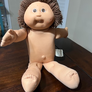 May include: A vintage Cabbage Patch Kid doll with a soft, flesh-toned body and a brown yarn hairstyle. The doll has a round face with rosy cheeks, large eyes, and a slightly open mouth. Its arms and legs are outstretched.
