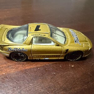 May include: A gold-coloured toy Firebird race car with the number 1 on the roof and side. The car has black wheels with Goodyear tyres and the words "True Value" on the side. The car is on a wooden surface.