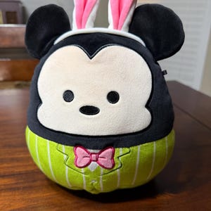 May include: A soft toy of a black and white character with large black ears and a white face. It is wearing pink bunny ears and a green and white striped outfit with a pink bow tie. The toy is on a wooden surface.