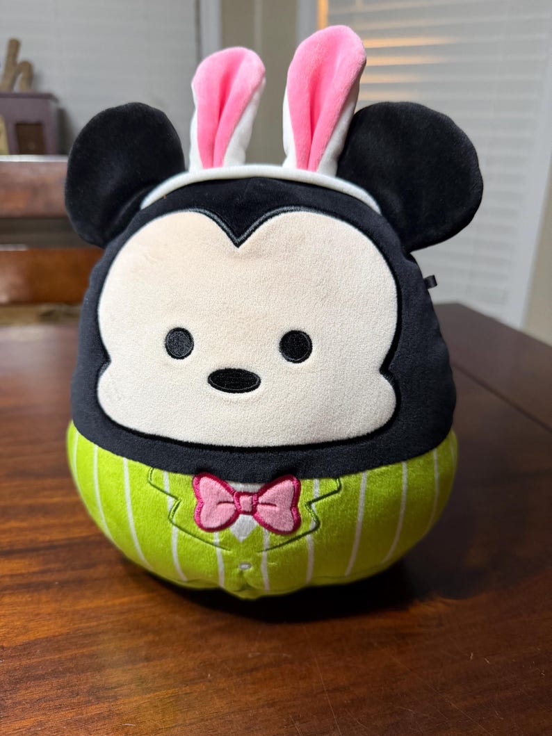 May include: A soft toy of a black and white character with large black ears and a white face. It is wearing pink bunny ears and a green and white striped outfit with a pink bow tie. The toy is on a wooden surface.