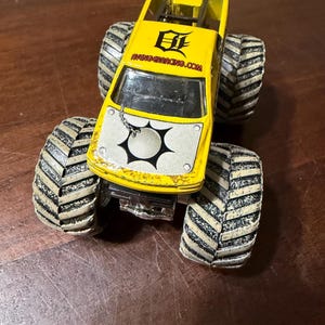Puede incluir: Un cami&oacute;n monstruo de juguete amarillo con neum&aacute;ticos grandes en blanco y negro. El cami&oacute;n tiene un panel frontal blanco con un dise&ntilde;o en blanco y negro y el texto "AVENGERACING.COM" en el lateral.