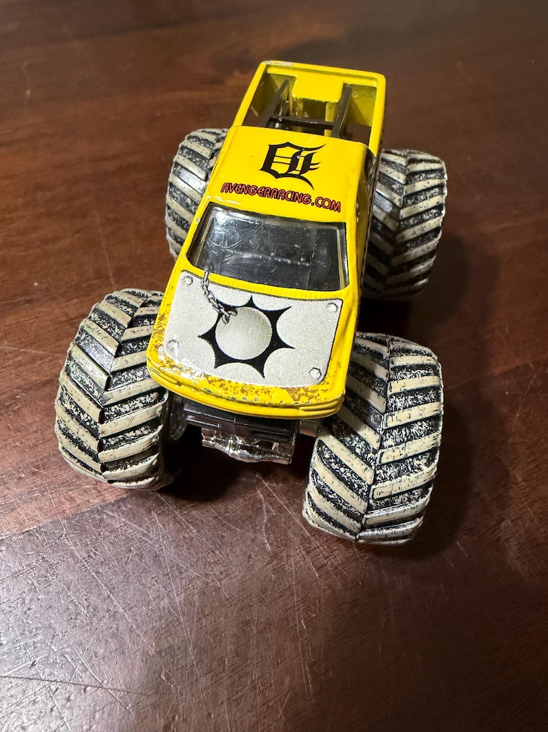 Puede incluir: Un cami&oacute;n monstruo de juguete amarillo con neum&aacute;ticos grandes en blanco y negro. El cami&oacute;n tiene un panel frontal blanco con un dise&ntilde;o en blanco y negro y el texto "AVENGERACING.COM" en el lateral.