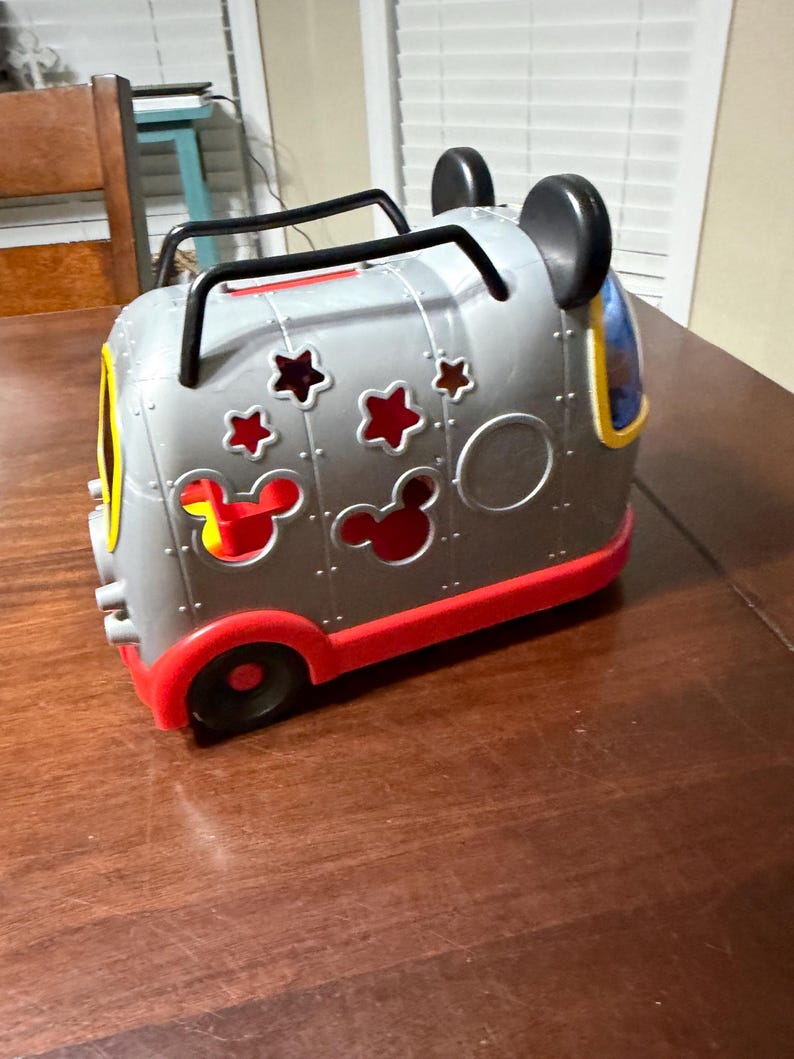 K&ouml;nnte beinhalten: Ein graues und rotes Spielzeugfahrzeug mit schwarzen Griffen und Mickey Mouse Ohren. Das Fahrzeug hat Stern- und Mickey Mouse-Kopf-Ausschnitte sowie gelbe und blaue Akzente. Die R&auml;der sind schwarz und rot.